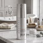 A tall silver pump bottle of SKIN-RG ENGLAND Cellular Balance Toner stands on a white marble bathroom counter, with a bathtub, folded towels, soap bars, and a small jar of cream blurred in the background.