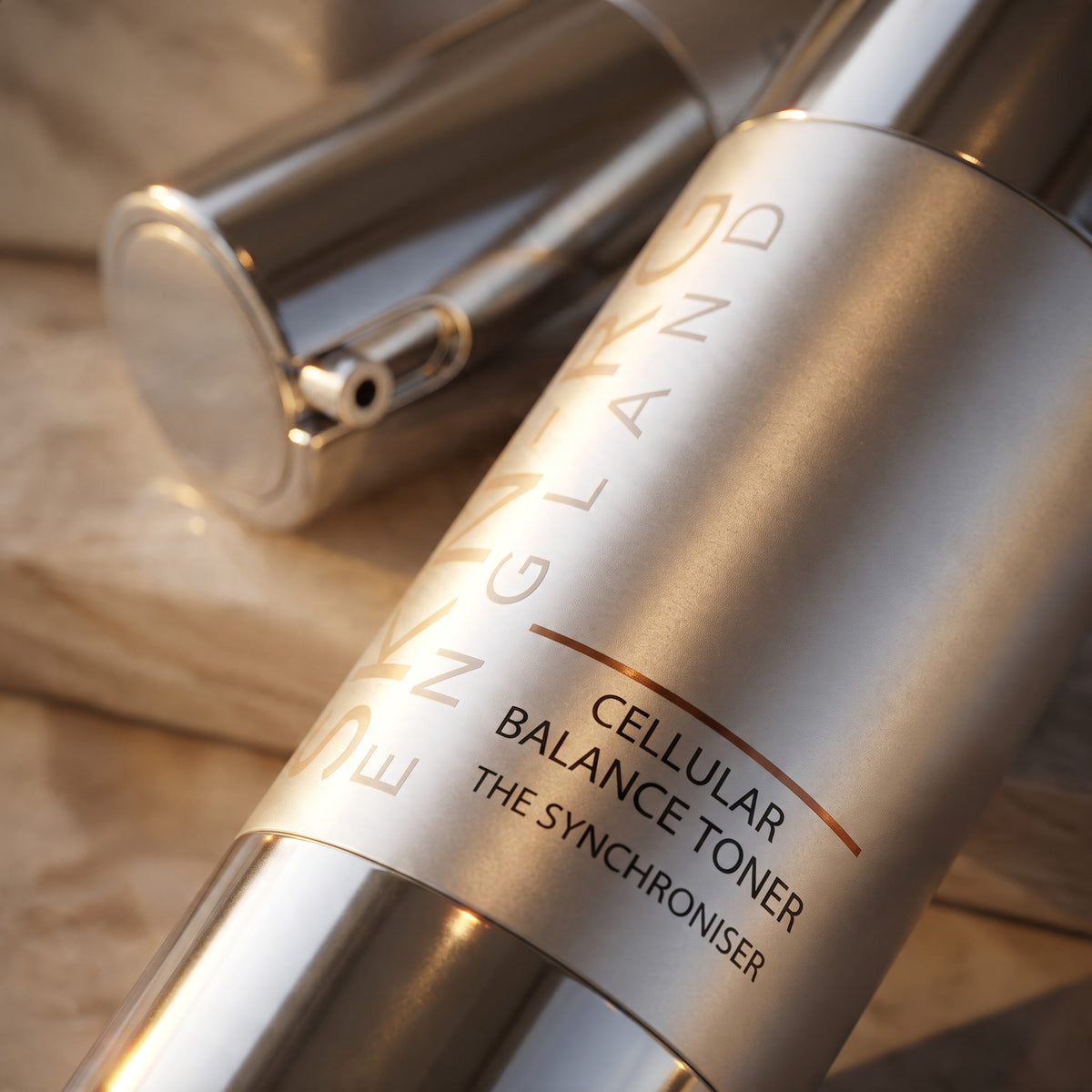 A close-up of a metallic silver skincare bottle labeled 'SKIN-RG ENGLAND Cellular Balance Toner  on a soft beige fabric background.