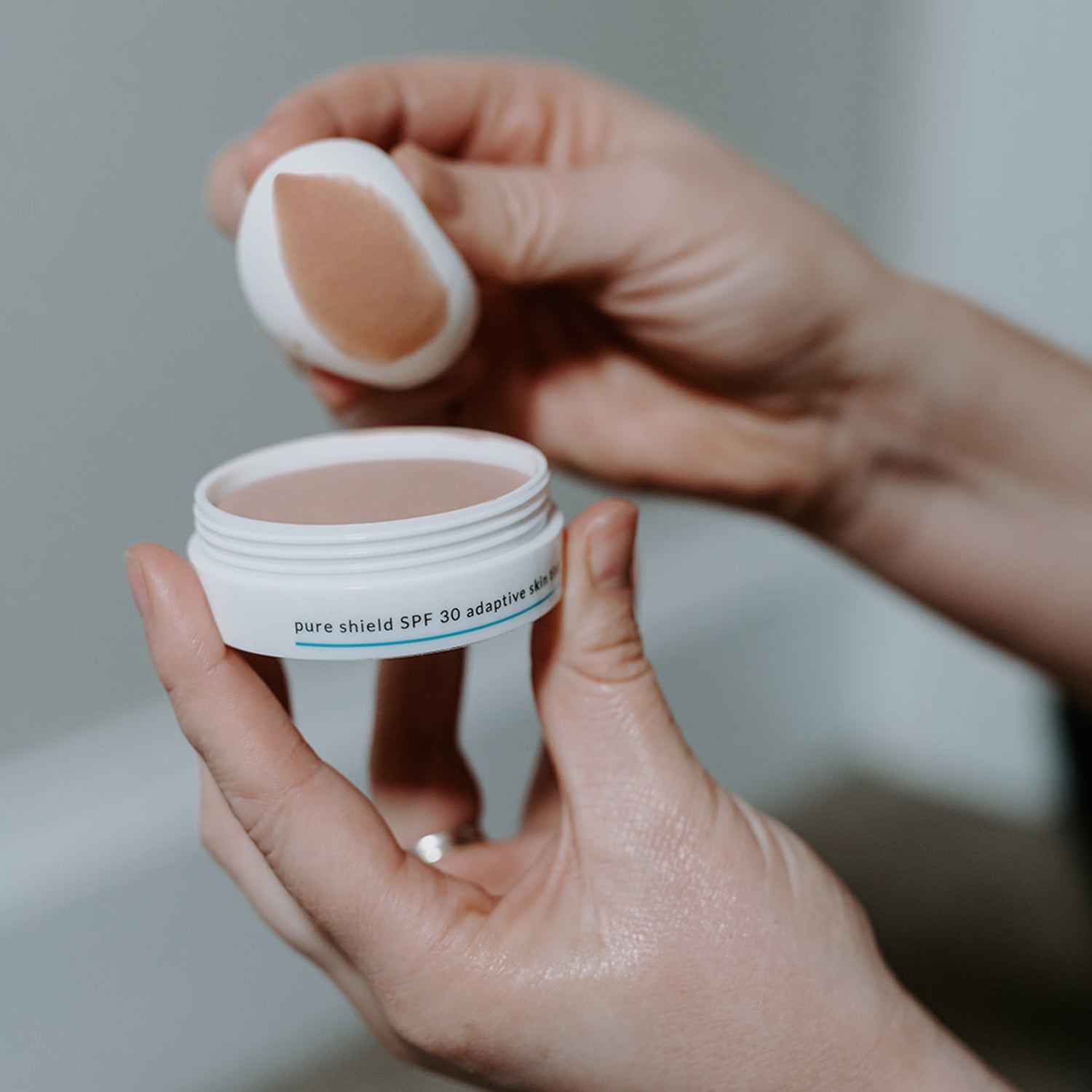 White jar with Pure Shield SPF 30 Adaptive Skin Tint with 'SKIN-RG' branding and words Pure Shield held in hands with applicator showing colour and texture of product. 