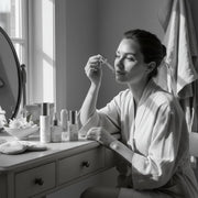 www.skin-rg.com woman applying serum from dropper to face, skincare bottles on vanity